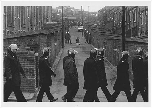 The greetings card oshowing a young woman returning home at lunchtime from her job at Hintons, the grocers. Police line the alleyway down which she is walking and a line of marching police officers pass the end of the alleyway.